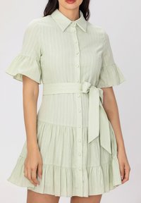Robe verte clair à manches courtes avec un col, boutonnière à l'avant et ourlet volanté. Présente une taille ceinturée assortie et un tissu texturé.