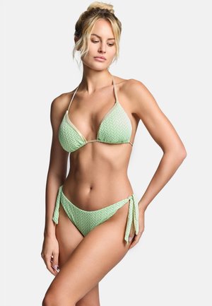 Frau mit blondem Haar, die einen hellgrünen, strukturierten Bikini mit seitlichen Bindebändern trägt, posiert mit einer Hand an der Hüfte vor einfarbigem Hintergrund.