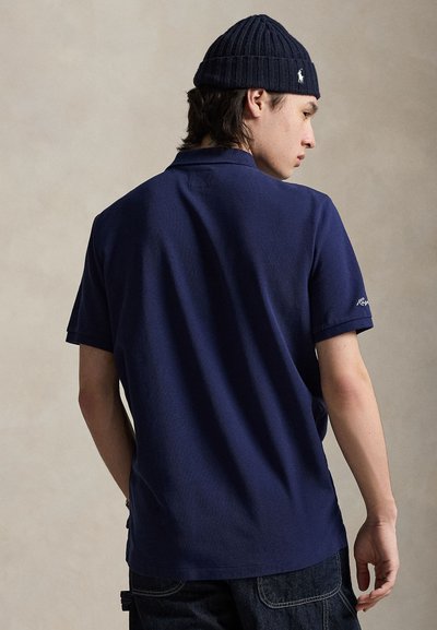 Tumesinine polo-särk lühikeste varrukatega ja kraega. Omaduseks on tekstureeritud kangas ja paremal varrukal tikitud logo. Kandmisel tumedad teksapüksid.