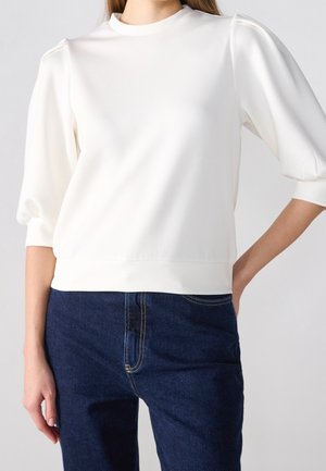 Vrouw die een witte blouse met pofmouwen van 3/4 lengte en een hoge hals draagt, gecombineerd met donkerblauwe spijkerbroek tegen een effen achtergrond.
