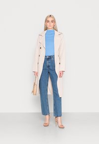 Helle beige Trenchcoat mit großen Knöpfen, ein blauer Rollkragenpullover, hochtaillierte blaue Jeans und nudefarbene Absatzsandalen, kombiniert mit einer kleinen beige Tasche.