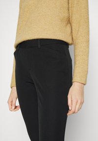 Black slim-fit trousers with a smooth texture, featuring side pockets and a fitted waistband, paired with a light tan knit sweater.