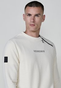 Sudadera blanca acolchada con cuello redondo. Presenta un zip lateral y un logotipo negro en el pecho, así como un parche en la manga. Tejido texturizado.