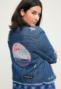 Chaqueta de mezclilla con un gran diseño bordado circular de palmeras y un atardecer, con detalles desgastados y un parche que dice "LA VIDA ENTERA".