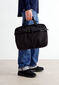 Sort stof laptop tas med to frontlommer, to topgreb og en struktureret rektangulær form. Båret af en person med blå jeans og sorte sko.