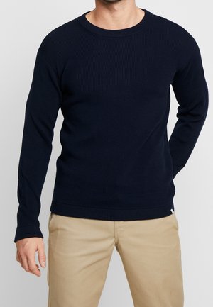 Homme portant un pull ajusté côtelé à manches longues bleu marine et un pantalon beige, debout avec une main derrière le dos.