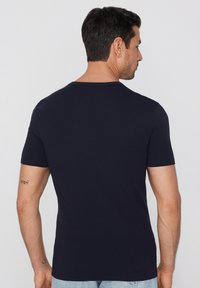 Mężczyzna o krótkich ciemnych włosach, ubrany w gładki granatowy t-shirt z krótkim rękawem, zwrócony tyłem na tle jasnoszarym.