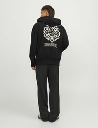 Hanorac negru cu glugă, având un design grafic floral pe spate, asociat cu pantaloni negri largi și sandale.