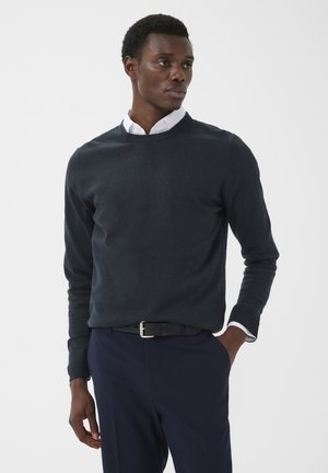 Mann trägt einen dunklen Pullover über einem weißen Hemd mit Kragen und marineblaue Hose, steht mit einer Hand in der Tasche und schaut zur Seite.