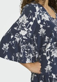 Robe florale marine avec imprimé botanique blanc. Avec des manches courtes en volants et un design en V. Fabriquée en tissu léger et texturé.