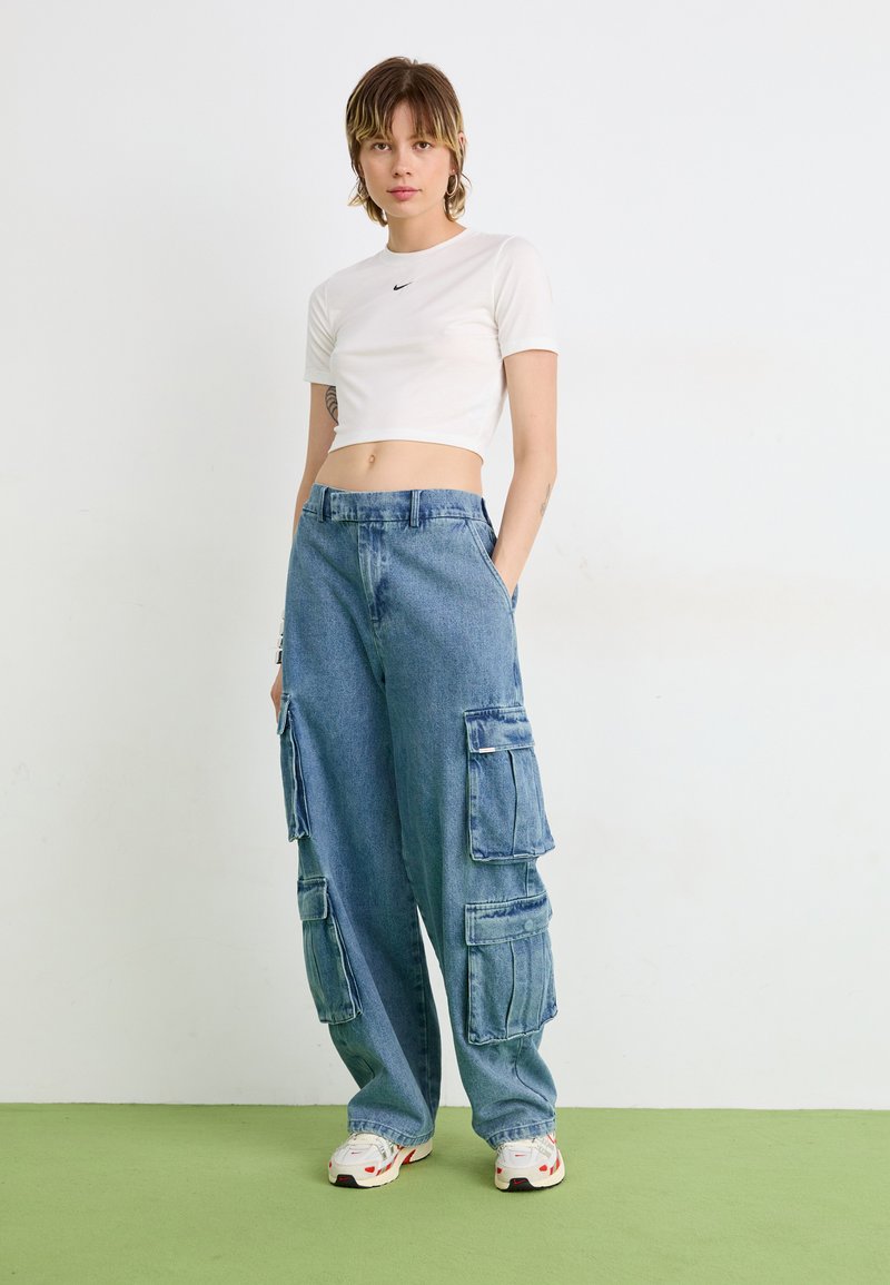 Weißes, zugeschnittenes T-Shirt mit kleinem Logo, kombiniert mit weit geschnittenen blauen Denim-Cargo-Hosen, die mehrere Seitentaschen und eine lockere Passform aufweisen.