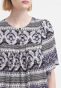 Blusa a motivi in bianco e nero con dettagli viola, caratterizzata da un collo rotondo, maniche corte e dettagli plissettati sul petto.