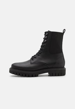 Sort læderkampstøvle med tyk rillede sål, snørelukning foran, trækkehank ved hælen og elastisk sidepanel, vist i sideprofil.