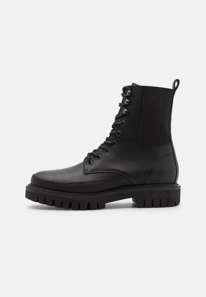 Botte de combat noire en cuir avec une semelle épaisse à crampons, lacets à l'avant, languette au talon et panneau latéral élastique, vue de profil.