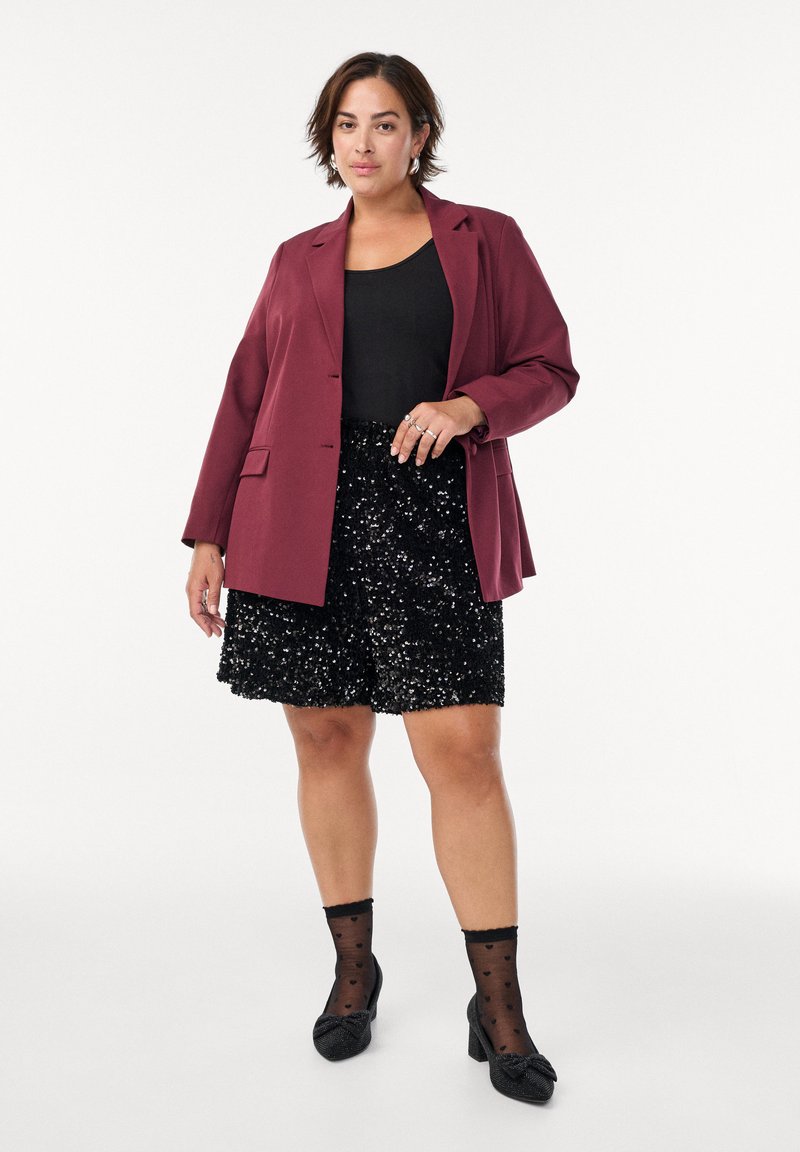 Blazer bordeaux su una canotta nera, abbinato a pantaloni corti neri con paillettes. Calze nere a pois e scarpe nere decorate completano l'outfit.