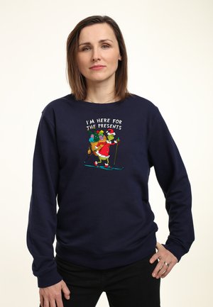 Marinblå sweatshirt i bomullsblandning med en färgglad illustration av Grinchen som åker skidor med presenter, texten ovan lyder "JAG ÄR HÄR FÖR PRESENTEN."