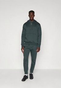 Sweatshirt com capuz verde escuro com bolso frontal, cordão no capuz e detalhe de logótipo. Combinado com calças de jogging combinando e sapatilhas desportivas pretas.