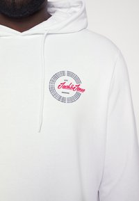 Bílá mikina s kapucí vyrobená z měkkého materiálu. Obsahuje kulatý motiv s nápisem "Jack & Jones" v červené barvě a textem v černé. Nastavitelná šňůrka.