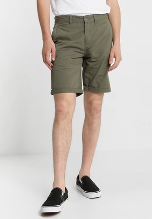 Homme portant un short vert olive à ourlet retroussé, un t-shirt blanc décontracté et des baskets noires sans lacets sur un fond clair uni.