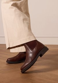 Bruine leren enkellaarzen met een gladde afwerking, elastische zijpanelen en een stevige zwarte zool, gecombineerd met lichtbeige broek met wijde pijpen.