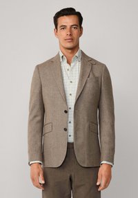 Brauner Tweed-Blazer mit Reverskragen, zwei vorderen Taschen und schwarzen Knöpfen, getragen über einem karierter Hemd. Glatte Textur und taillierte Passform.