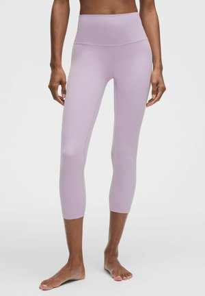 Kvinde iført lyse lilla højtaljede, korte leggings stående barfodet mod en ensfarvet baggrund.