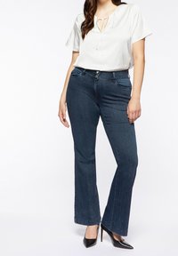 Witte blouse met een strikhalslijn, gecombineerd met donkerblauwe flared jeans. De jeans hebben vijf zakken en zijn gemaakt van denimstof met een gladde textuur.