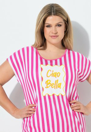 Donna con capelli biondi che indossa una maglietta a righe rosa e bianca con la scritta "Ciao Bella", in piedi con le mani sui fianchi su sfondo bianco.