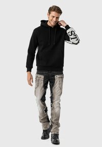 Zwarte hoodie met witte zijaccenten en tekst. Draagt het met versleten, lichtgrijze jeans met zwarte vlekken en scheuren. Zwarte laarzen.