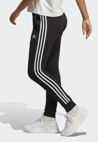 Melnas sporta bikses, kas izgatavotas no mīksta auduma, ar trim baltām svītrām gar sāniem un Adidas logo uz kreisā augšstilba.