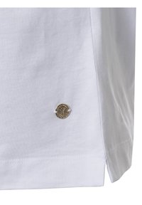 Witte stof met een klein ronden metalen embleem met een boomdesign, genaaid nabij de onderzoom van een kledingstuk.