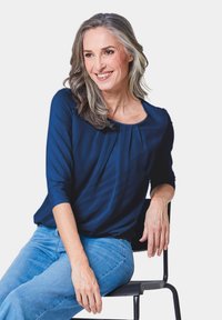 Marineblaues Oberteil mit gefältelten Details, 3/4-Ärmel, kombiniert mit hellblauen Jeans. Model sitzt auf einem schwarzen Stuhl vor hellen Hintergrund.