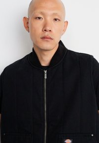 Črna izolirana usnjena brezrokavnik z zadrgo, okroglim ovratnikom in dvema sprednjima žepoma, izdelana iz trpežnega materiala, z nekim majhnim logotipom na žepu.