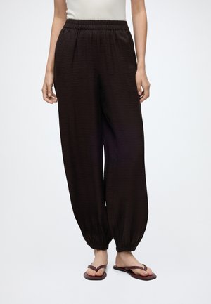 Femme portant un pantalon noir texturé ample avec taille et poignets élastiques, associé à des tongs marron foncé, debout sur fond blanc.