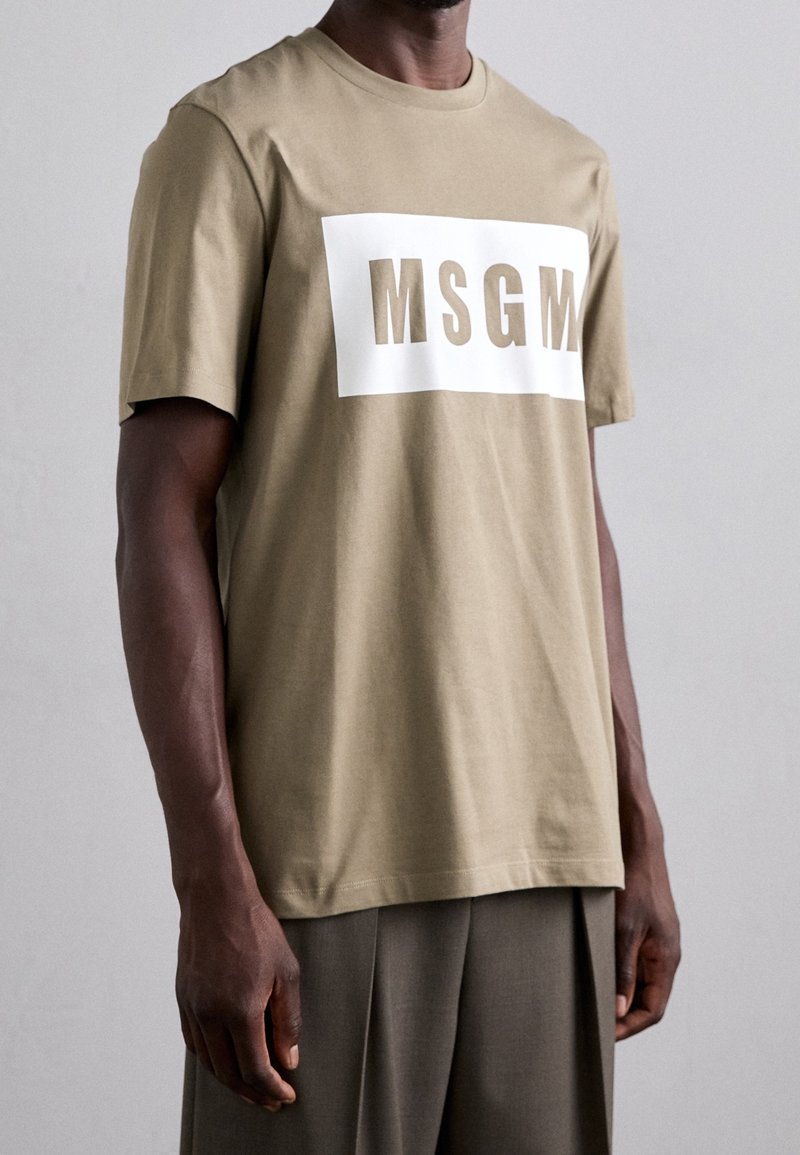 T-shirt en coton beige avec un graphique rectangulaire blanc portant l'inscription "MSGM." Manches courtes et col rond. Texture douce, coupe décontractée.