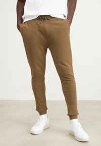 Bruine joggerbroeken gemaakt van zacht materiaal met een trekkoord in de taille, taps toelopende pijpen en twee zijzakken, gecombineerd met witte sneakers.