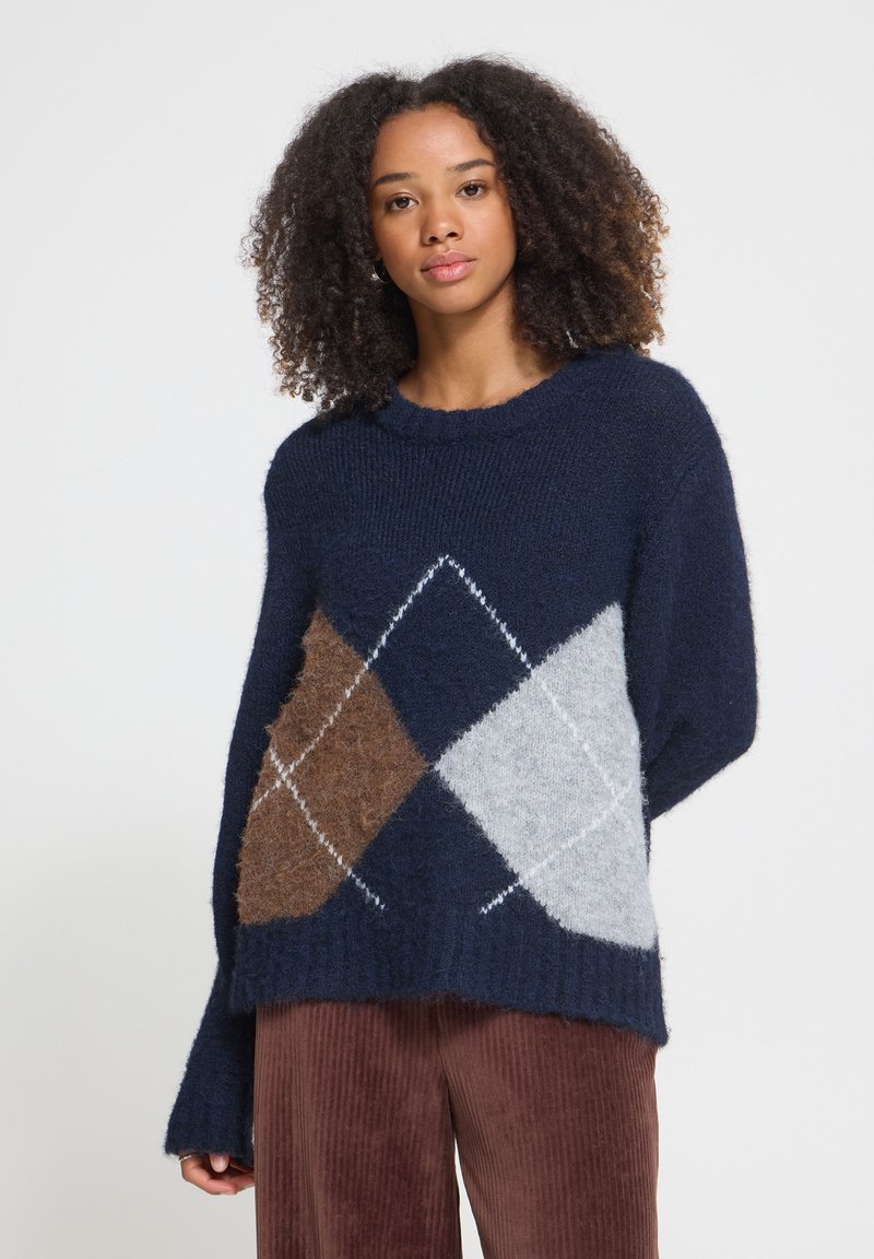Jeune femme aux cheveux bouclés portant un pull bleu marine avec des motifs losanges marron et gris, et un pantalon en velours marron.