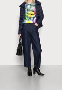 Personne portant un pull rayé coloré, une doudoune sombre, un jean large foncé, des bottines à talons noires, tenant un sac à main noir, debout sur un sol gris.