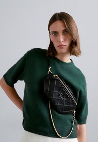 Geantă din piele matlasată neagră, tip crossbody, cu curea din lanț și închidere cu fermoar, având un emblema rotundă pe față. Pulover tricotat verde.
