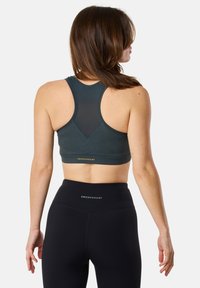 Mörkgrön sport-bh med racerback-design och meshnätspanel, med guld-logotyp. Matchas med svarta högmidjade leggings.