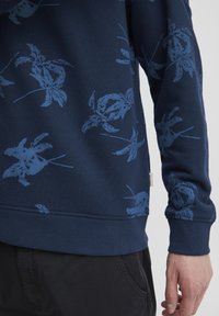 Mørkeblå hettegenser med blå blomstertryk, ribbestrikkede mansjetter og nederkant, myk tekstur, og en liten logo-lapp på siden.