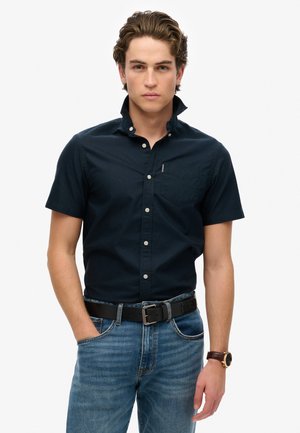 Camisa de botones de manga corta en color azul marino con cuello, bolsillo y botones blancos; se lleva con unos vaqueros de denim azul claro y un cinturón negro.