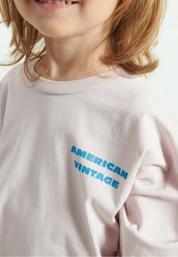 Svijetlo ružičasta majica kratkih rukava s rebrastim izrezom. Na lijevoj strani ima natpis "AMERICAN VINTAGE" ispisan odvažnim plavim slovima. Izrađena od mekanog pamuka.