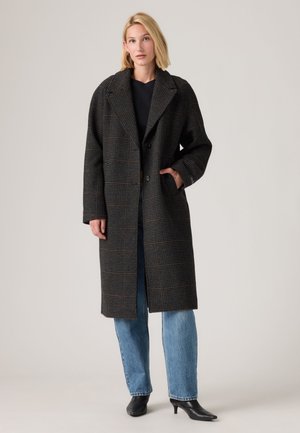 Femme aux cheveux blonds portant un long manteau foncé à carreaux, un haut noir, un jean bleu et des bottines noires à talons, debout devant un fond uni.
