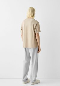 Person med axellångt blont hår står med ryggen mot oss iförd en beige t-shirt med kort ärm, ljusgrå byxor och vita sneakers.