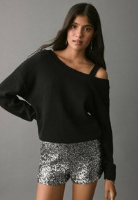 Czarny sweter bez ramiączek z teksturowanej tkaniny, w zestawieniu z błyszczącymi, srebrnymi szortami pokrytymi cekinami, z dopasowanym krojem i elastyczną talią.