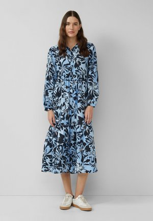 Robe à manches longues, boutonnée en bleu clair avec un imprimé floral bleu foncé. Jupe mi-longue évasée avec taille élastique et tissu texturé. Portée avec des baskets blanches.
