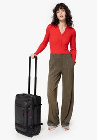 Black wheeled suitcase with two handles and red zipper pulls. Model wears a red long-sleeve buttoned top and olive green wide-leg pants.
