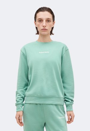 Άτομο που φοράει φούτερ Björn Borg σε μέντα πράσινο και παντελόνια που ταιριάζουν, στέκεται μπροστά σε μια απλή ανοιχτογκρι φόντο.