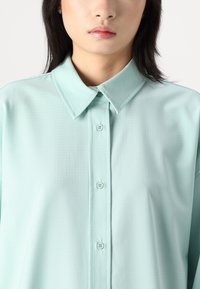 Camicia button-up verde menta chiaro con colletto strutturato, tessuto liscio con un sottile motivo a griglia e sei bottoni sulla parte anteriore.